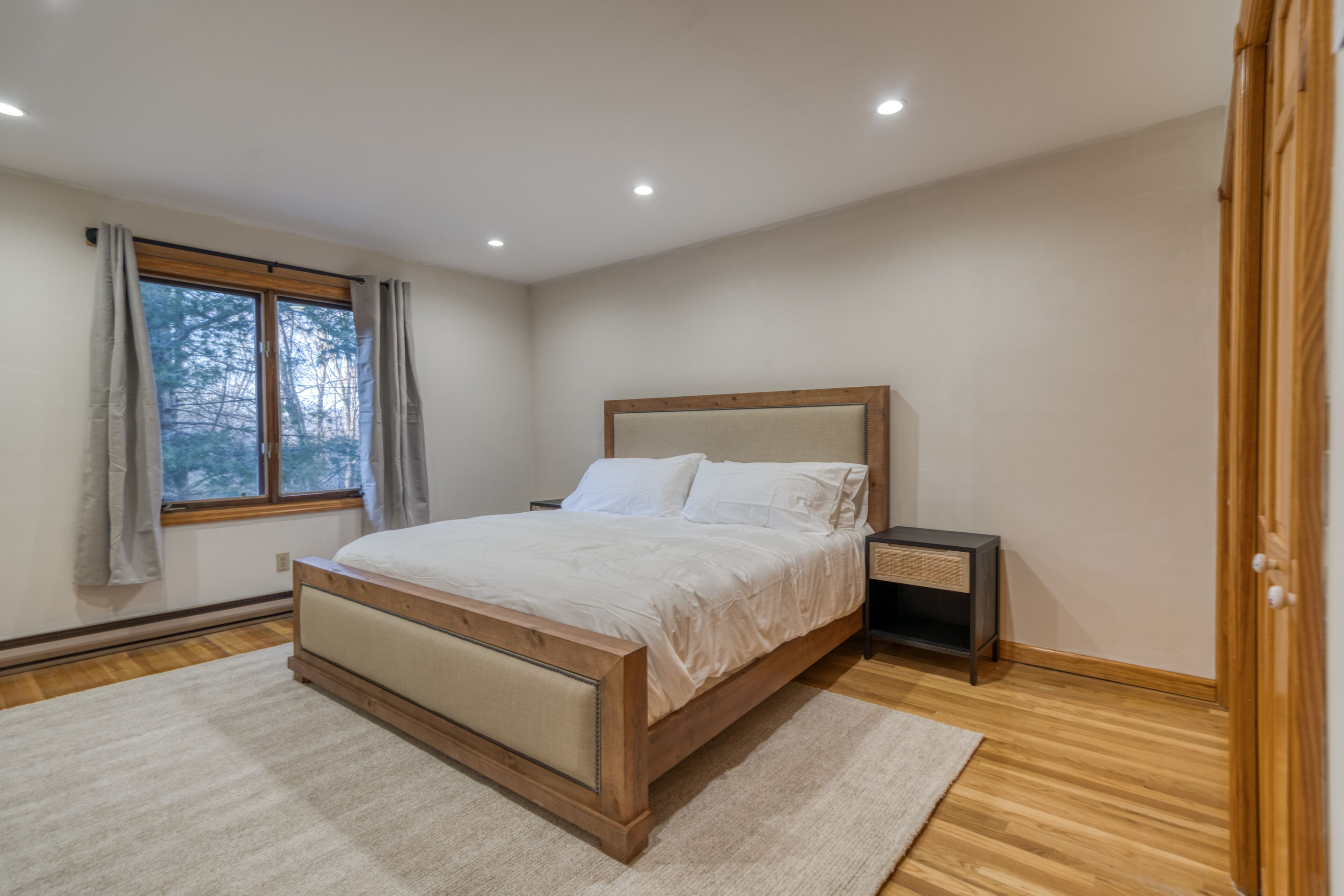 A bedroom with a wooden bed, a nightstand, a window with curtains, and a view of trees outside.