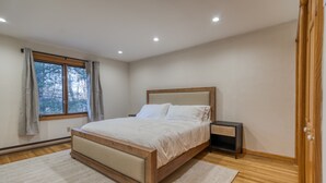 Ein Schlafzimmer mit einem hölzernen Bett, einem Nachttisch, einem Fenster mit Vorhängen und Blick auf Bäume.
