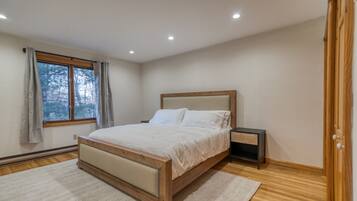 A bedroom with a wooden bed, a nightstand, a window with curtains, and a view of trees outside.