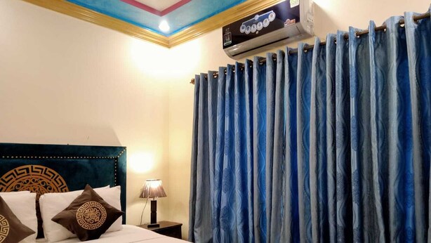 A hotel room with a bed, a nightstand with a lamp, and a ceiling-mounted air conditioner. - Infinity Hotel  (Lahore)