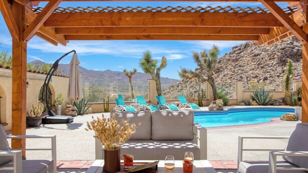 Ein Poolbereich mit einer Holzpergola, Liegestühlen und einem Tisch mit Getränken und einer Vase mit Trockenblumen.