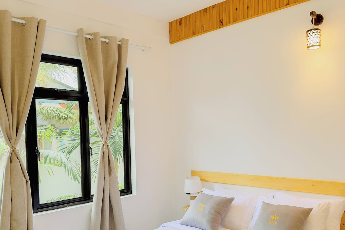 A neatly made bed with white linens and a wooden headboard, a window with curtains, and a wall-mounted light fixture.