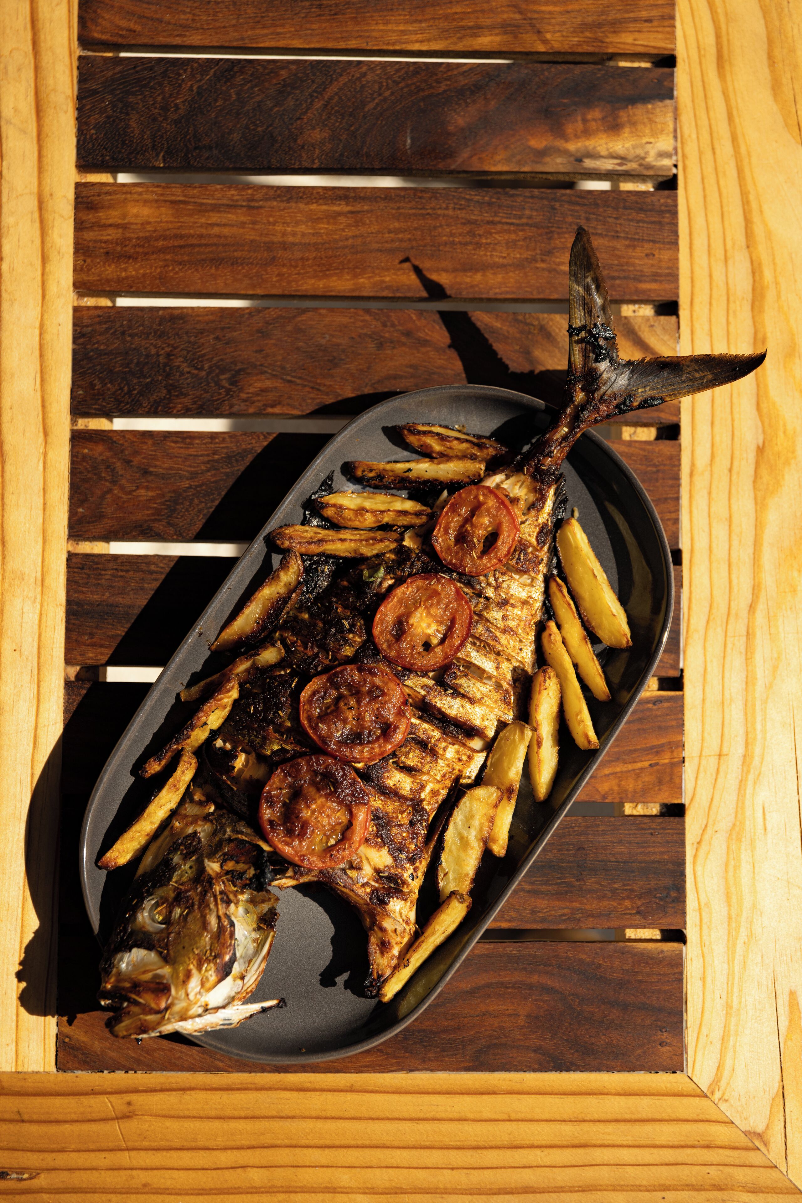 A fish with grilled potatoes and tomatoes on a black plate on a wooden table.