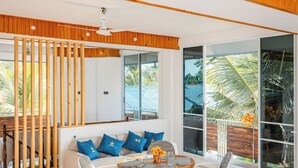 A modern living room with a wooden ceiling, a white sofa with blue cushions, a dining table with chairs, and large windows with a view of palm trees. - Dhoani Maldives Guesthouse (Kendhoo)