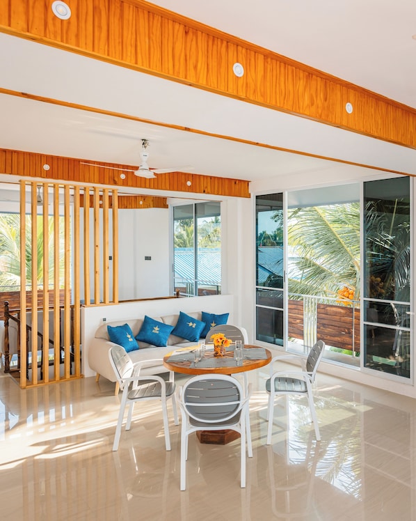Una sala de estar moderna con techo de madera, un sofá blanco con cojines azules, una mesa de comedor con sillas y ventanas amplias con vista a palmeras.