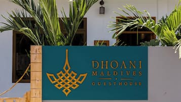 Placa indicando Dhoani Maldives Guesthouse, com cerca de madeira e plantas de palmeira.