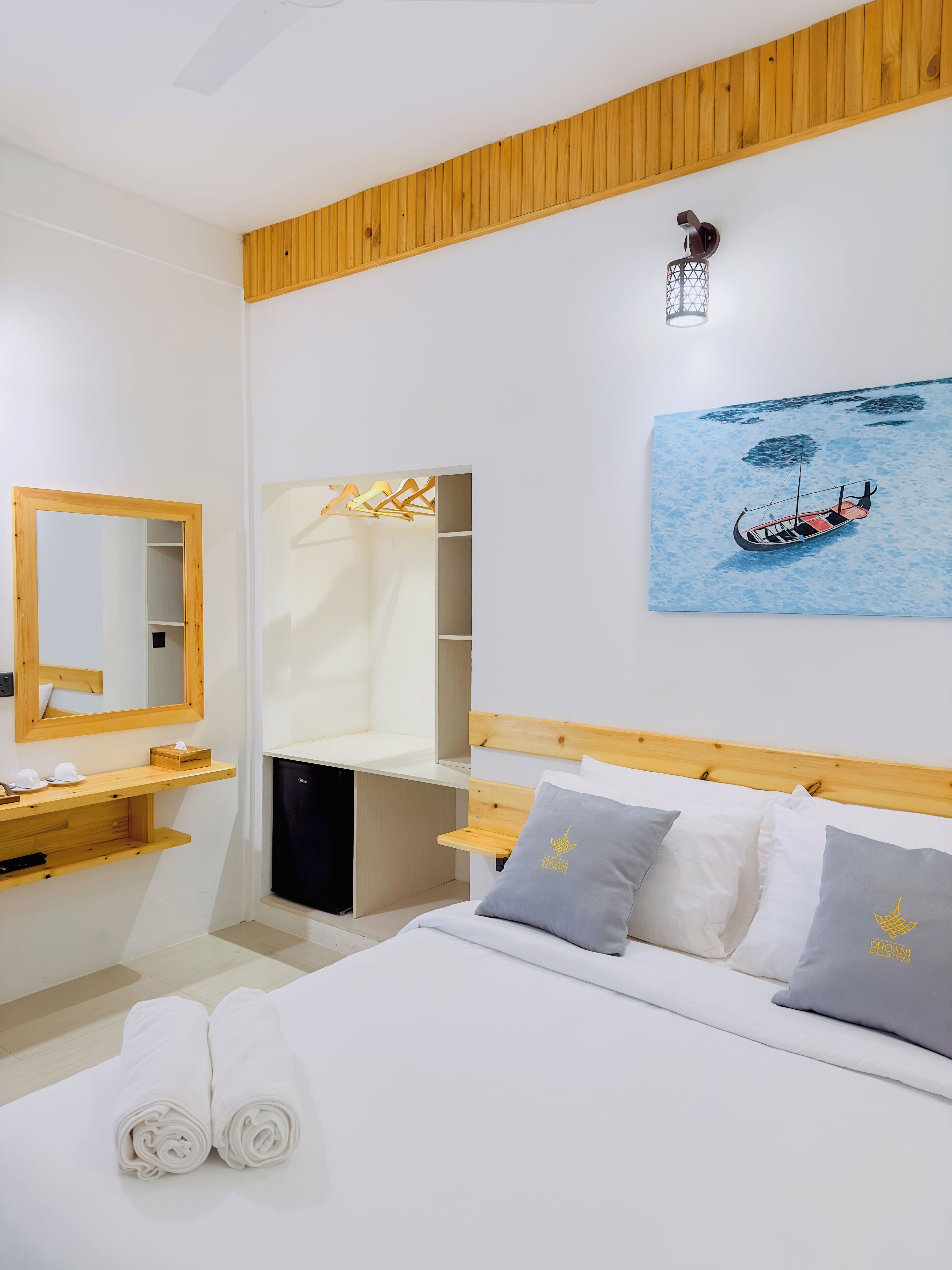 A hotel room with a bed, a wooden headboard, a bedside table with a mirror, a wardrobe, and a painting of a boat on the wall.