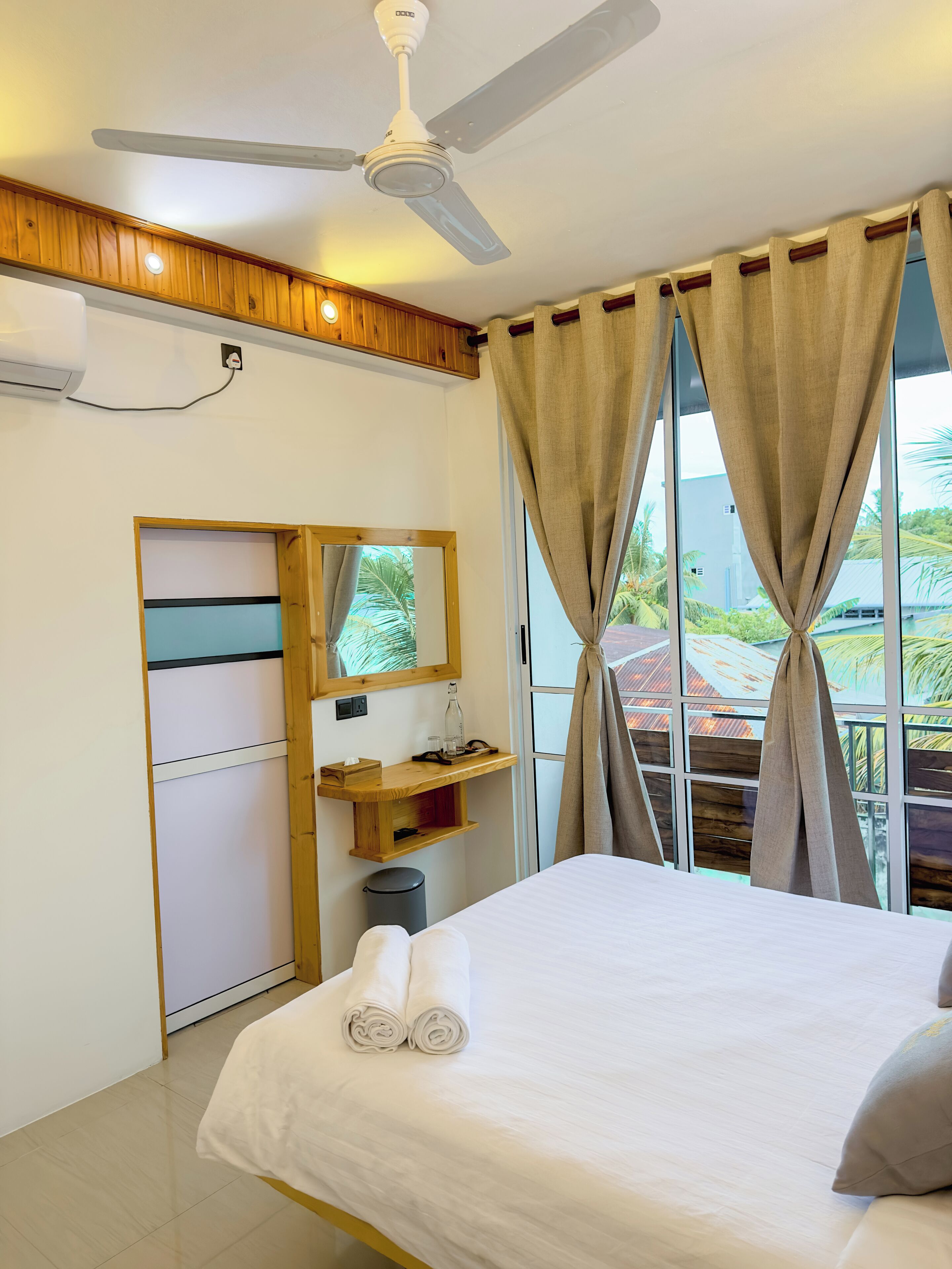 A hotel room with a bed, a ceiling fan, a window with curtains, a wooden shelf, and a mirror.