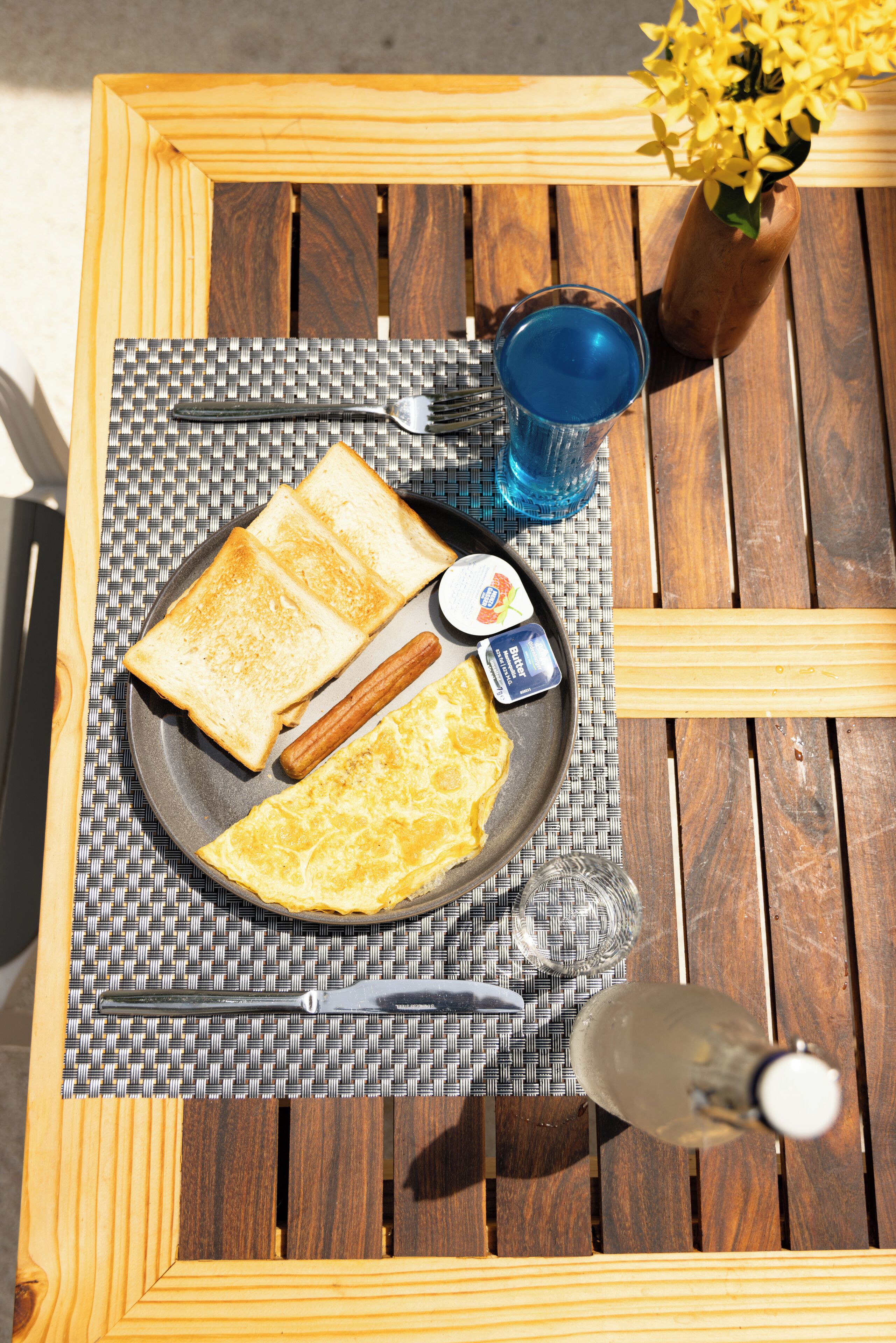 A wooden table with a woven placemat, a plate with toast, a hot dog, a glass of blue liquid, a bottle, and a vase with yellow flowers.