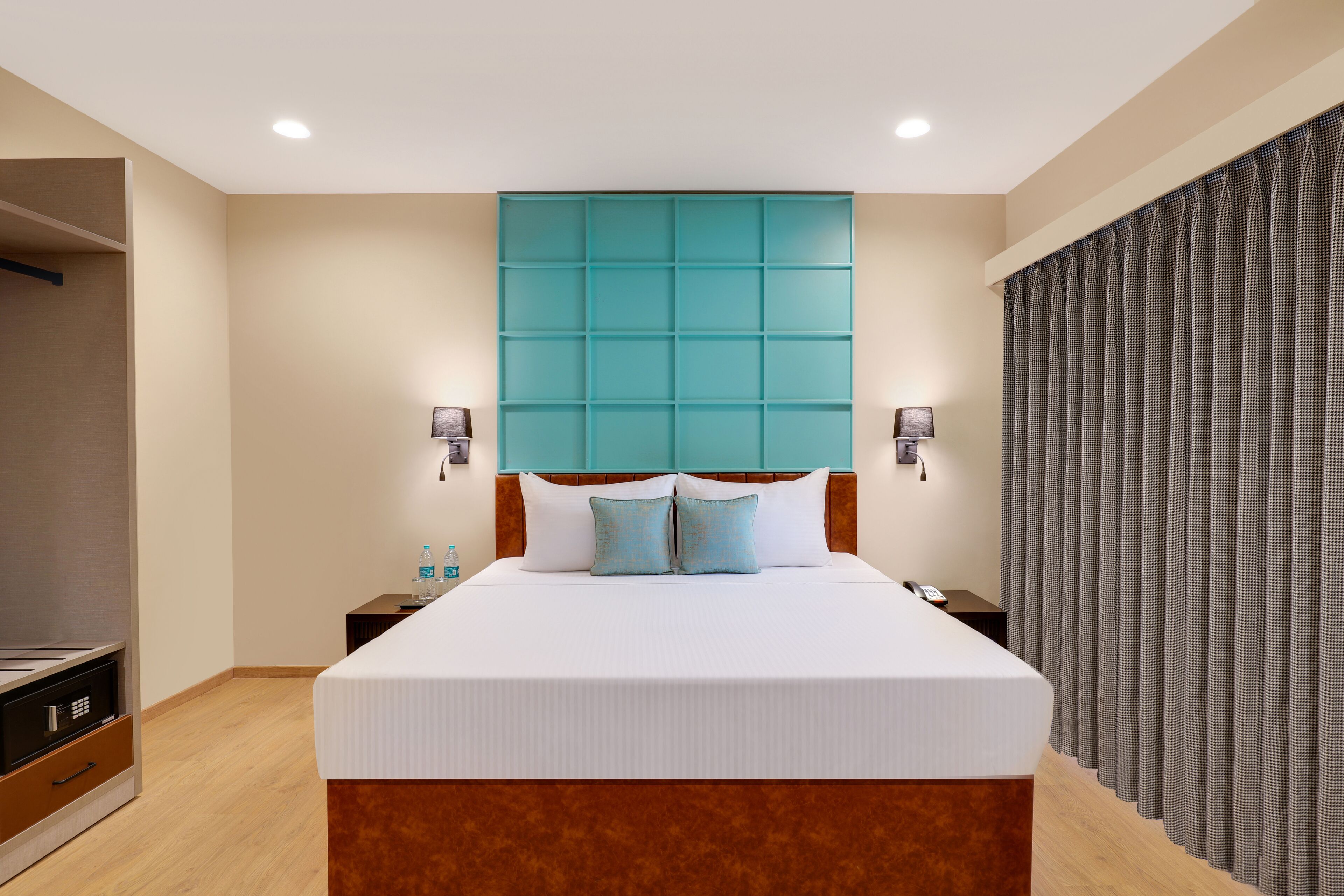 Una habitación de hotel con una cama grande, un cabecero de madera, dos mesitas de noche con lámparas y un amplio ventanal con cortinas.