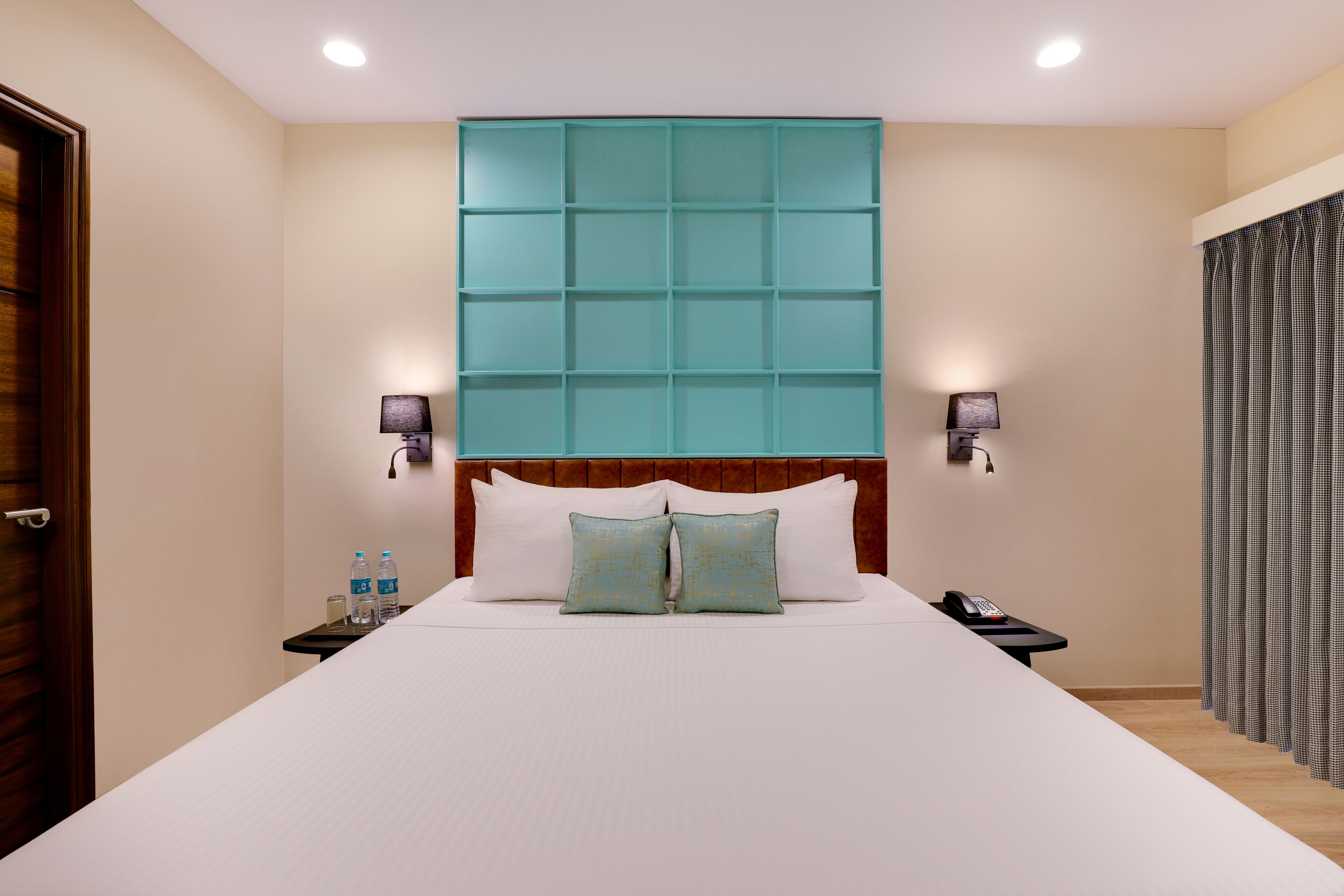 Una habitación de hotel con una cama grande, un cabecero con panel azul turquesa, dos lámparas de noche y una mesita de noche con un teléfono y botellas de agua.