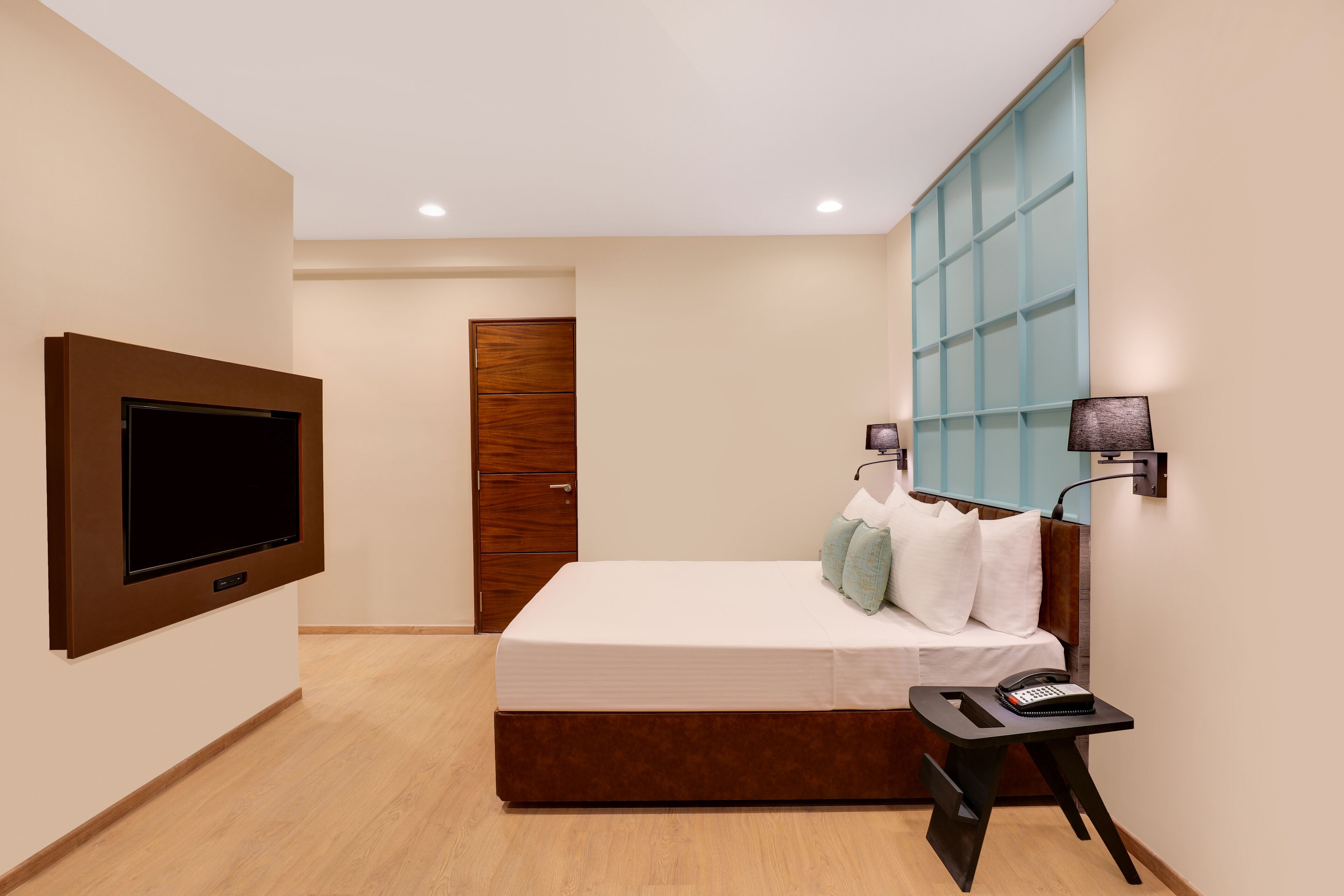 A modern hotel room with a large bed, a flat-screen TV mounted on the wall, a bedside table with a lamp, and a wooden wardrobe.