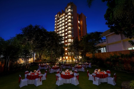 An outdoor banquet setup with round tables and white chairs, surrounded by trees and a building in the background.