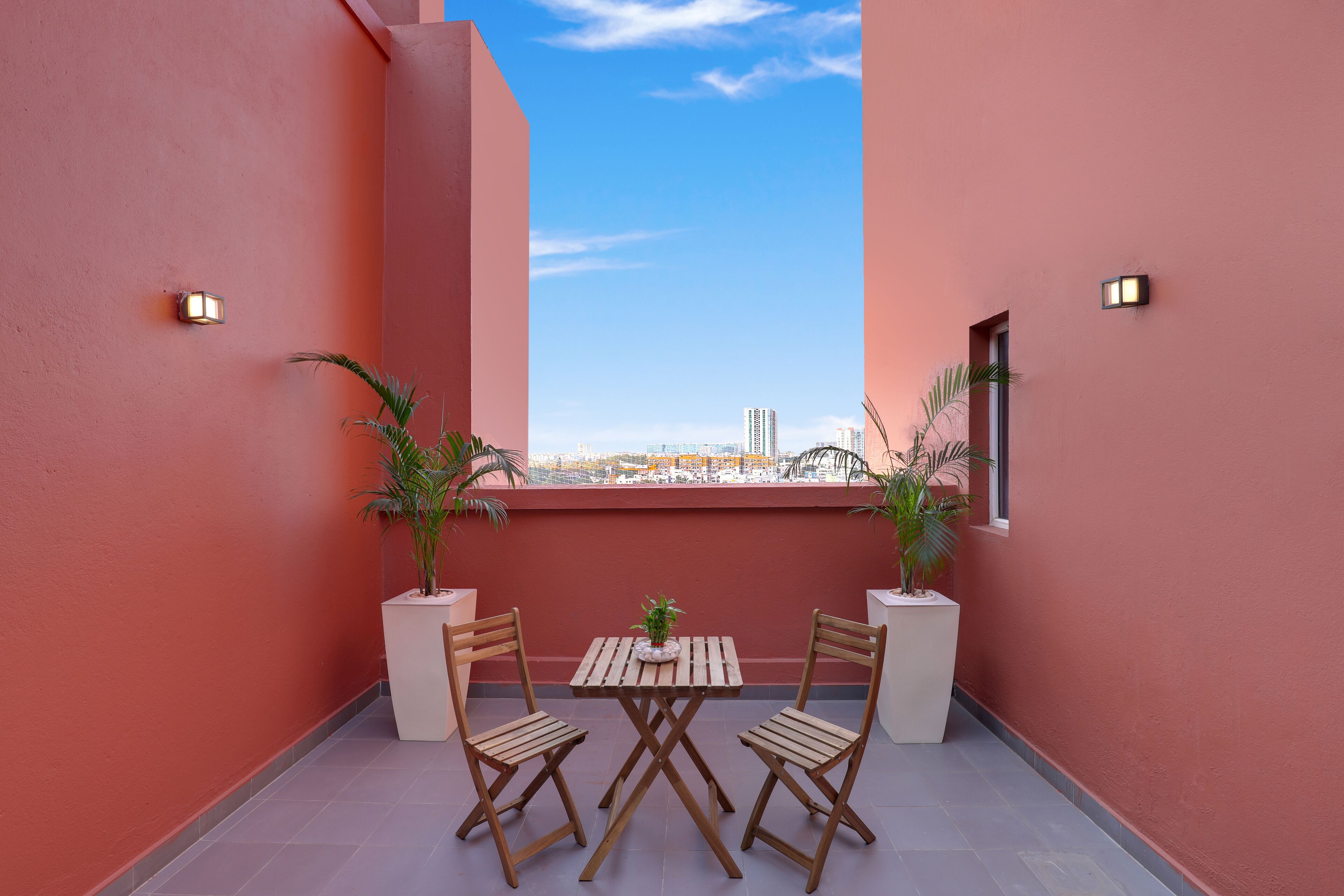 A balcony with two wooden chairs and a small table, potted plants, and a view of the cityscape.