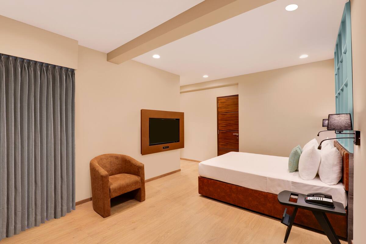 A modern hotel room with a bed, a TV mounted on the wall, a wooden wardrobe, a small armchair, and a desk with a telephone.