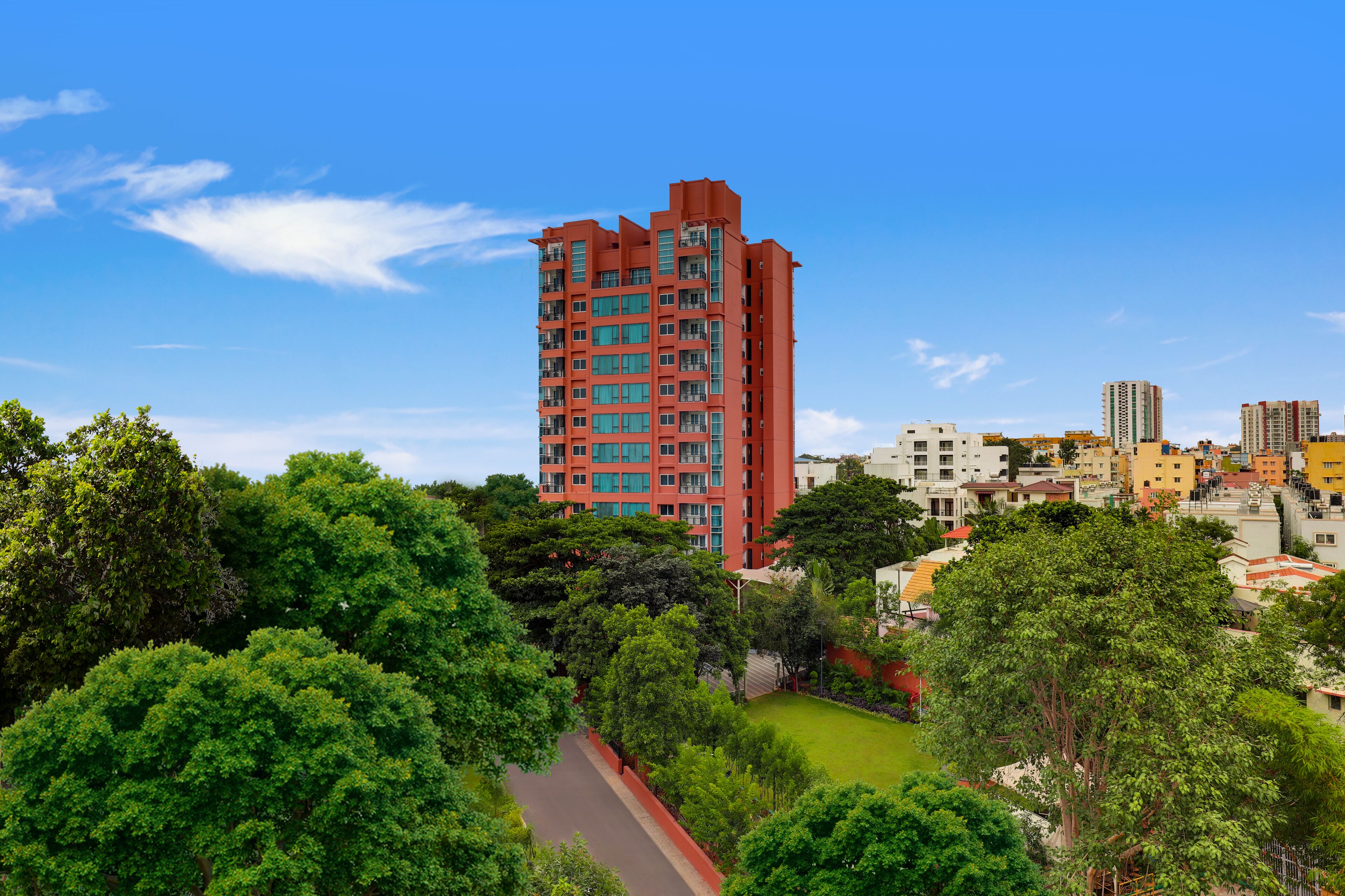 A high-rise building with multiple balconies, surrounded by lush green trees and a clear blue sky.