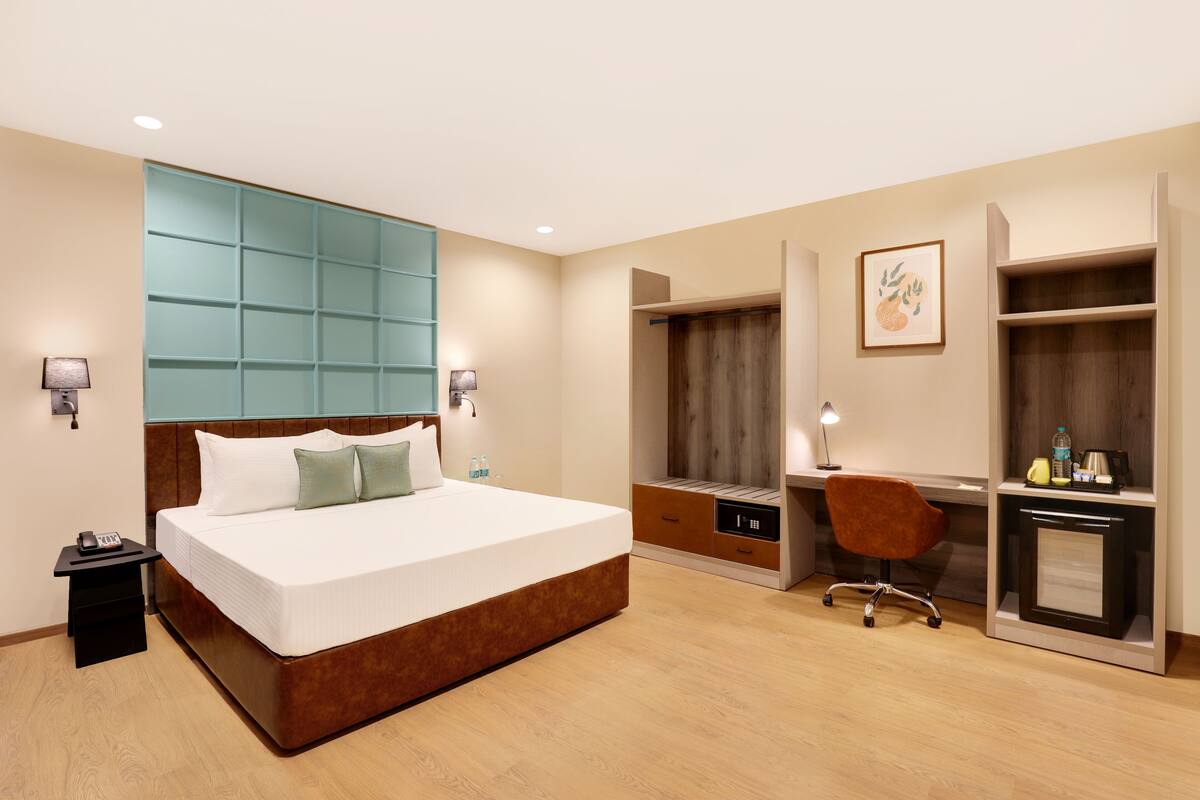 A hotel room with a large bed, a desk, a chair, a wardrobe, and a small table with a lamp.