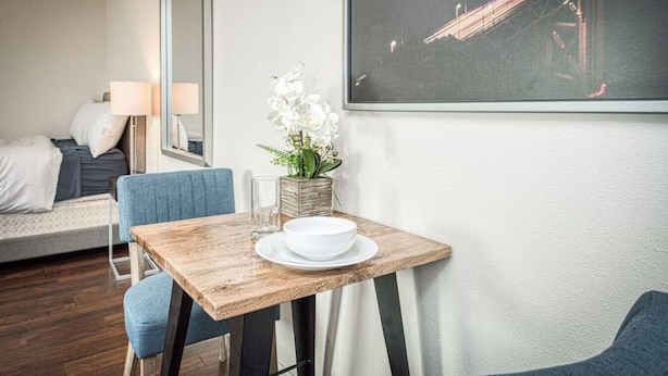 Une chambre d’hôtel avec une table à manger en bois, un lit avec une chaise bleue, une grande fenêtre donnant sur le paysage urbain et un vase avec des fleurs blanches.