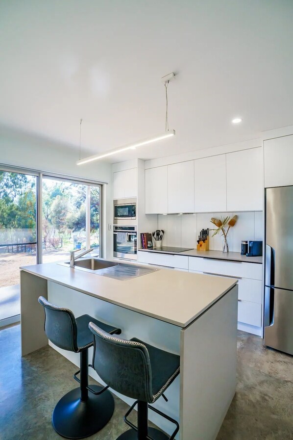 A modern kitchen with white cabinetry, a large island with bar stools, and a view of the outdoors. - Loxton Riverfront Apartment (Loxton)