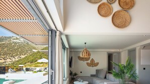 Ein modernes Wohnzimmer mit großem Fenster, das einen Pool und Hügel zeigt, eine Couch, zwei runde, weiße Tische und dekorative Wandbehänge.