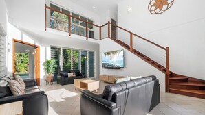 A spacious living area with a black leather sofa, a wooden coffee table, and a TV mounted on the wall. A wooden staircase with a railing leads to an upper level. - This stunning home is located in Palm Cove. Short 5min walk to the beach. (Palm Cove)