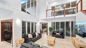 A spacious living area with a large black leather sofa, a wooden coffee table, and a TV mounted on the wall. There is a balcony with a wooden railing and large windows. - This stunning home is located in Palm Cove. Short 5min walk to the beach. (Palm Cove)