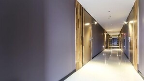 Un pasillo largo y estrecho con piso blanco brillante y paredes en tonos neutros. A ambos lados, el pasillo está bordeado de puertas, y luces en el techo proveen iluminación.