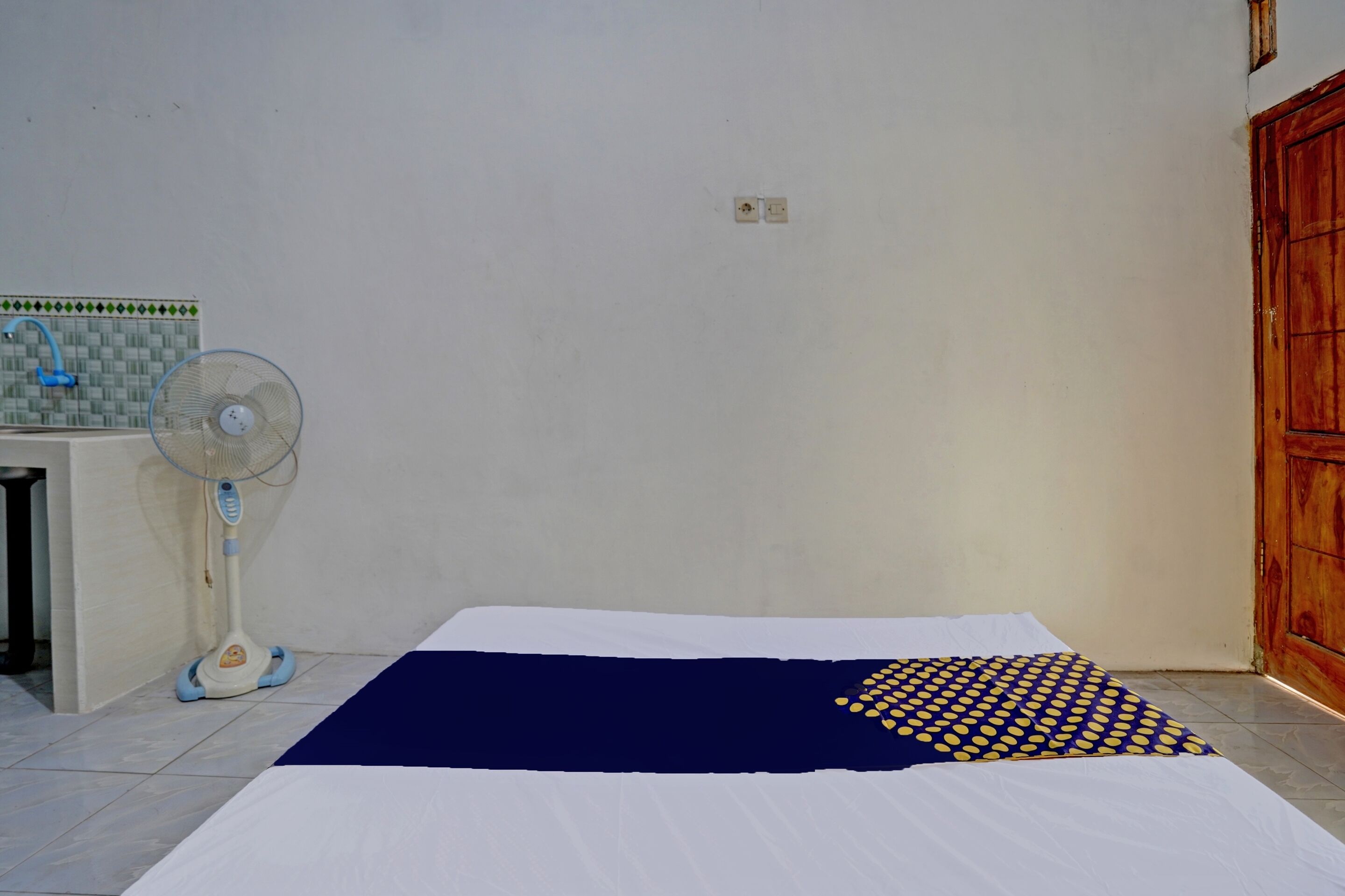 A room with a bed, a standing fan, a tiled wall, and a wooden door.