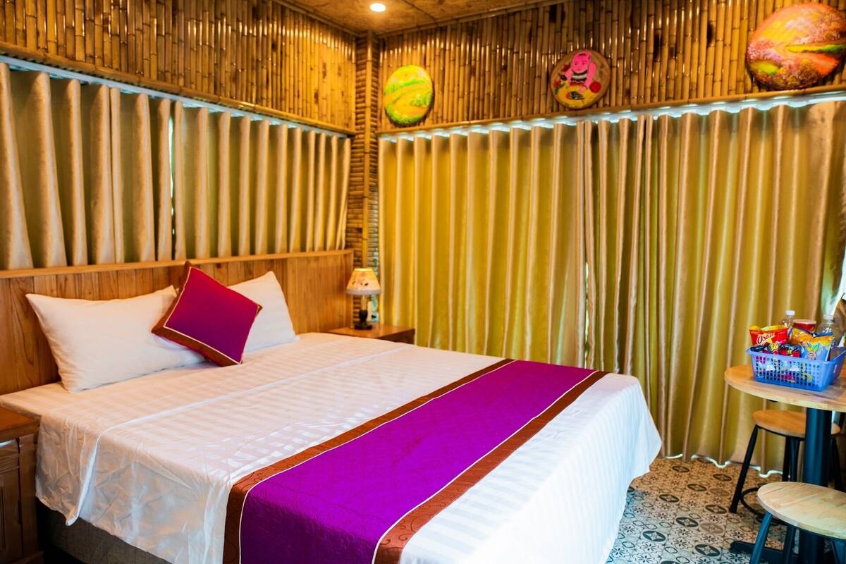A hotel room with a bed, a nightstand, a lamp, a small table with snacks, and a chair.