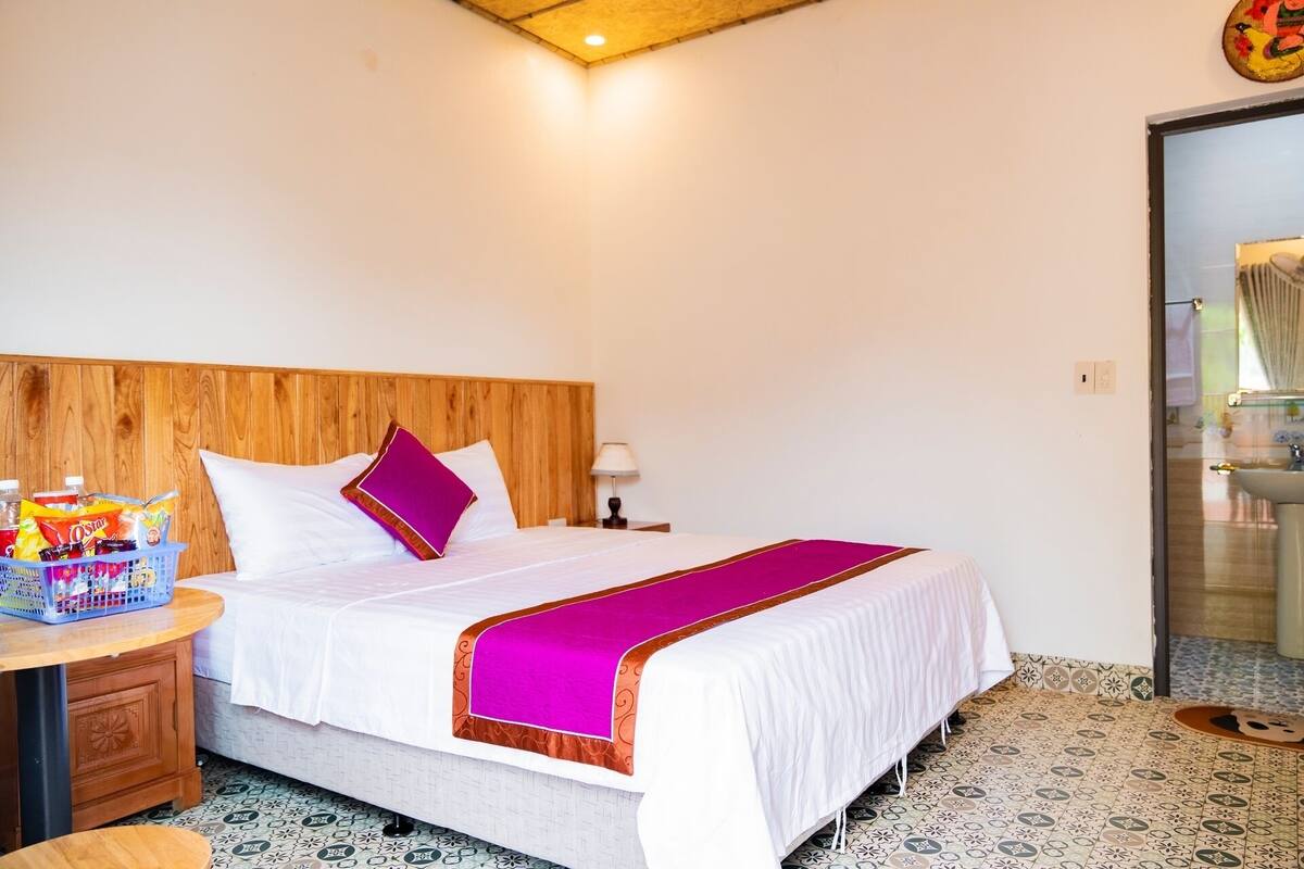 A hotel room with a wooden headboard, a bed with white bedding and a pink accent pillow, a small bedside table with a lamp, and a bathroom visible through an open door.