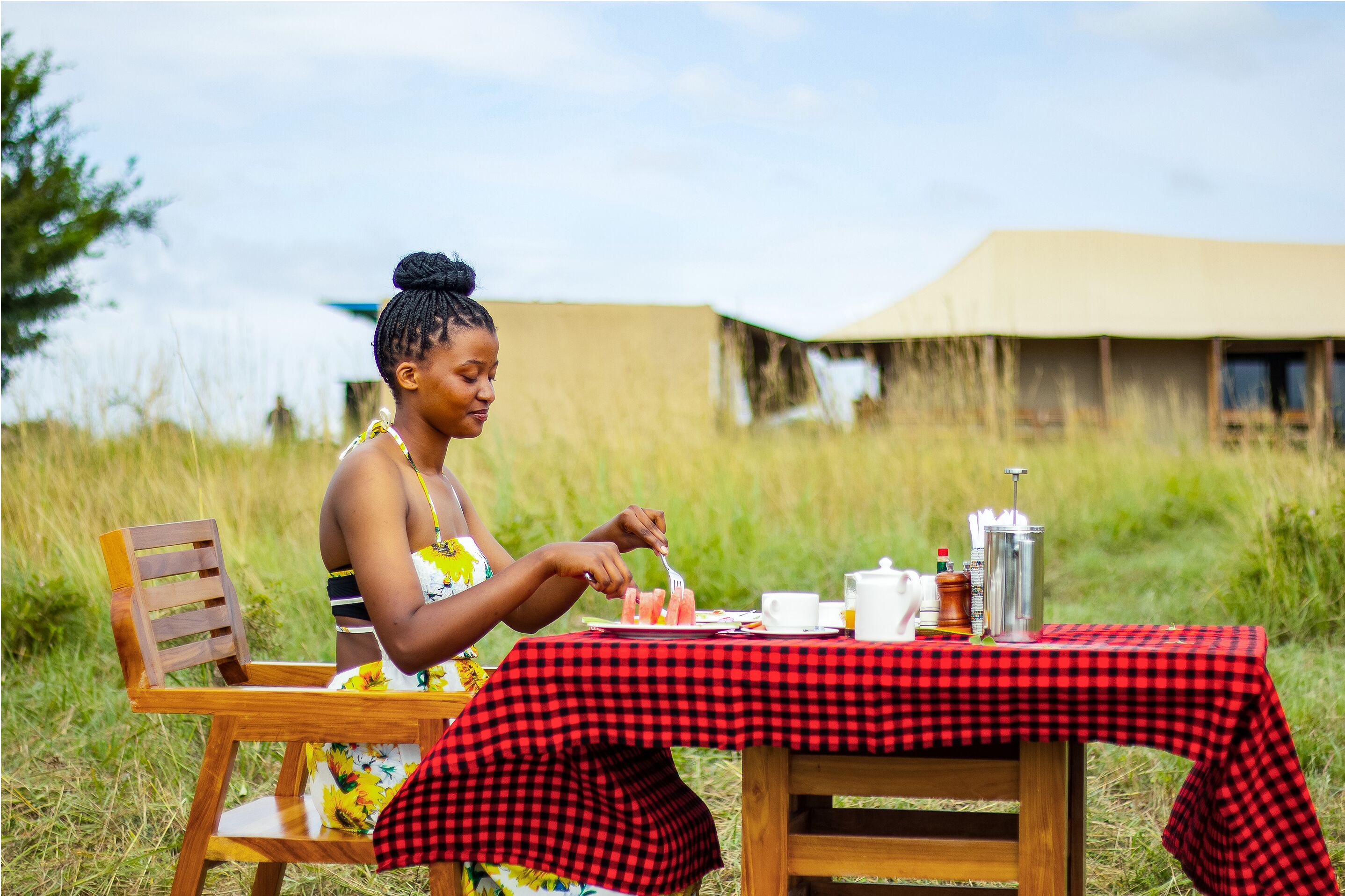 A person dining at an outdoor table with a checkered tablecloth, surrounded by a grassy field and a building in the background.