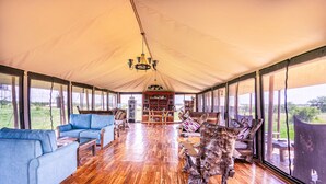 A tented accommodation with a wooden floor, blue sofas, a wooden coffee table, a bookshelf, and a large window offering a view of the outdoors. - Malaika Luxury Camp Seronera Serengeti (Serengeti)