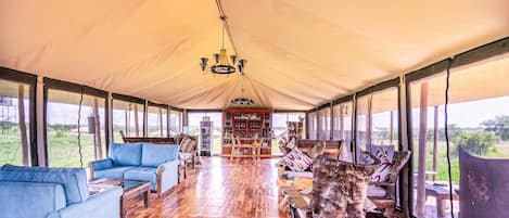 A tented accommodation with a wooden floor, blue sofas, a wooden coffee table, a bookshelf, and a large window offering a view of the outdoors.