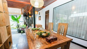 A wooden dining table set with food and drinks, surrounded by chairs, in a room with large windows and a wooden ceiling. - Gopala Villa North Ubud (Ubud)