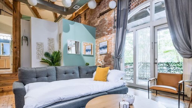 A modern living room with a grey sofa, a round wooden coffee table, and a view of a brick wall and greenery outside. - 100 Year-old Historic Brick 2BR Loft Large Patio2 (Durham)