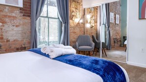 A hotel room with a brick wall, a large bed with blue bedding, a grey armchair, and a mirror reflecting the room. - 100 Year-old Historic Brick 2BR Loft Large Patio2 (Durham)