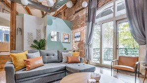 A modern living room with a grey sectional sofa, a round wooden coffee table, and a view of the outdoors through large windows. - 100 Year-old Historic Brick 2BR Loft Large Patio2 (Durham)