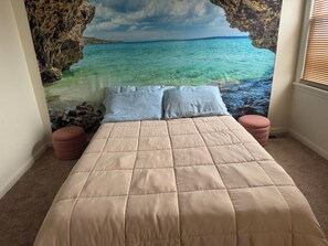 A bedroom with a bed, two pillows, a beige quilted bedspread, a rock wall mural, and a window with blinds. - Kennedy Melrose House (Atlantic City)