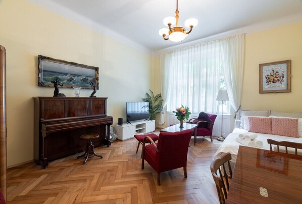 A living room with a wooden piano, a television, a dining table with chairs, a red armchair, a sofa, and a chandelier. - Historic Apartment close to Ludmila Cathedral by EasyBNB Premium (Prague)