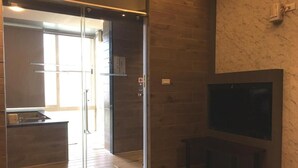 A modern interior with a glass door leading to a kitchen area, a flat-screen TV mounted on the wall, and a bench. - Now and Then Hot Spring Inn (Jiaoxi)