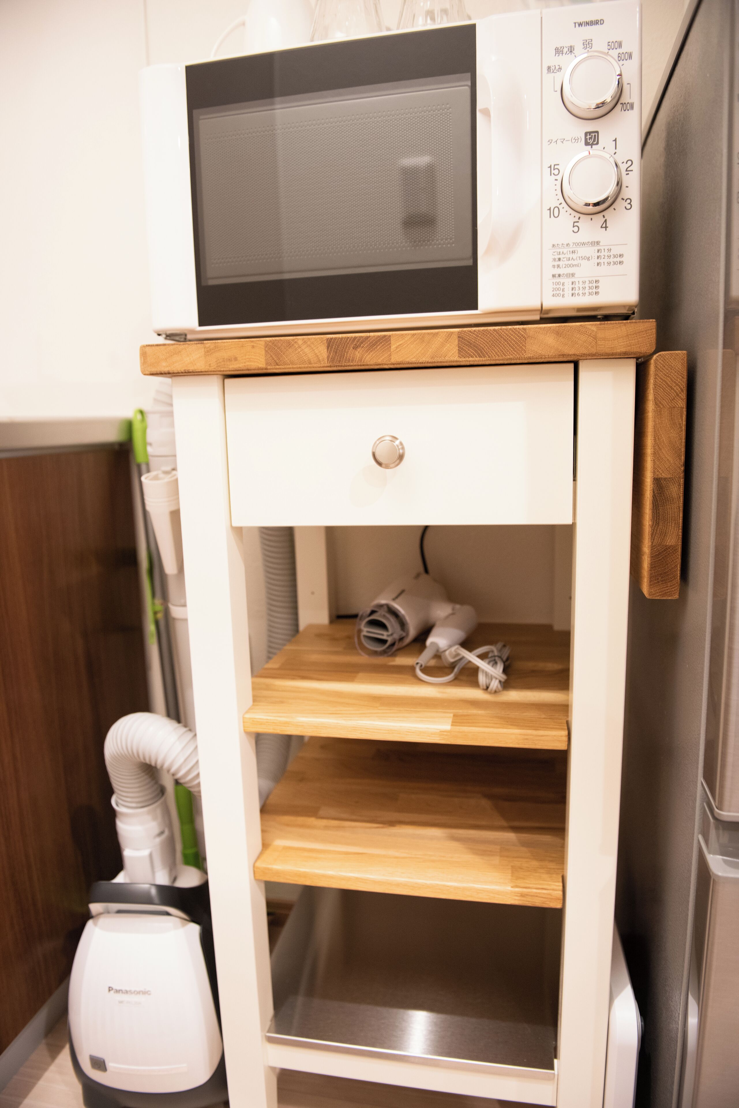 Une cuisine avec un four à micro-ondes posé sur une étagère en bois, un appareil de la marque Panasonic, et un meuble blanc.