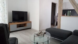 Ein modernes Wohnzimmer mit einem Flachbildfernseher auf einem Holzständer, grauen Sofas, einem Glastisch und einer Treppe, die zu einer oberen Ebene führt.