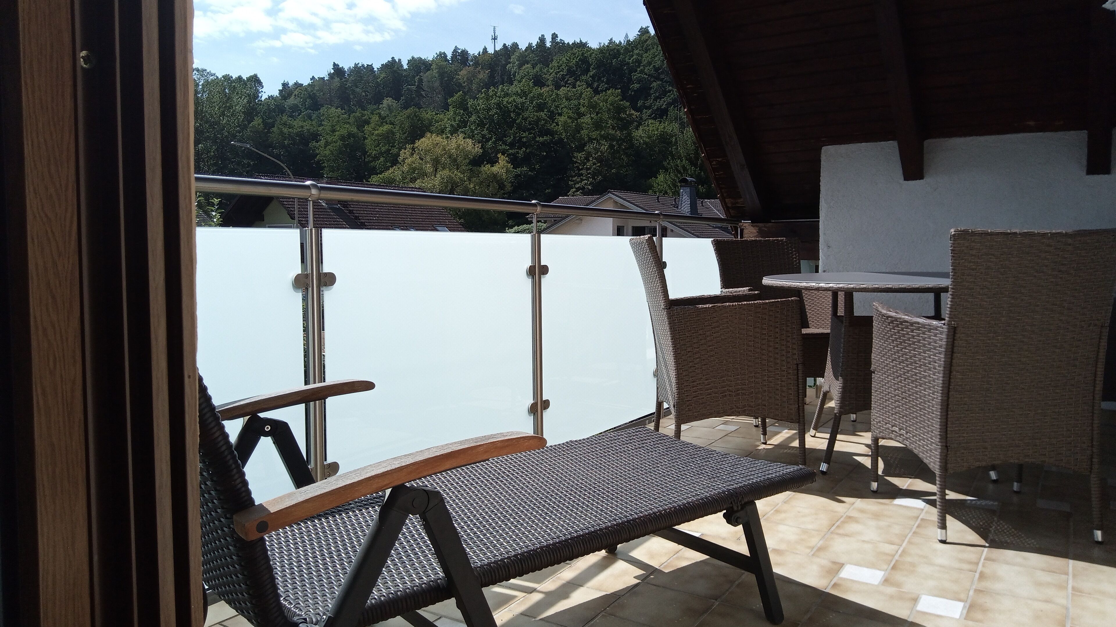 Varanda com cadeira de vime e uma pequena mesa, com vista para uma cerca branca e uma colina arborizada.