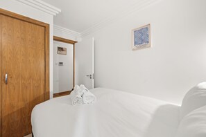 A hotel room with a wooden wardrobe, a bed with white linens, a framed artwork on the wall, and a door leading to another room.