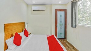 A bedroom with a wooden headboard, a bed with white and red bedding, an air conditioning unit, a door with a decorative glass panel, and a window with a view of trees. - Hotel Jeet Stay  (New Delhi)