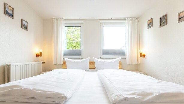 Un lit double avec du linge de lit blanc, dans une pièce dotée de deux fenêtres, de murs blancs et d’un radiateur.