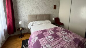 A bedroom with a bed, a red chair, a small table, and a wall with floral wallpaper. - Cozy 1BR Apartment with Balcony and Garage Near Beach and Downtown (Les Sables-d'Olonne)