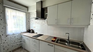 A modern kitchen with light grey cabinets, a stainless steel sink, and a window with patterned curtains. - Cozy 1BR Apartment with Balcony and Garage Near Beach and Downtown (Les Sables-d'Olonne)