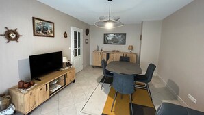 A compact living space with a TV, wooden entertainment unit, dining table with chairs, and a ceiling fan. - Cozy 1BR Apartment with Balcony and Garage Near Beach and Downtown (Les Sables-d'Olonne)
