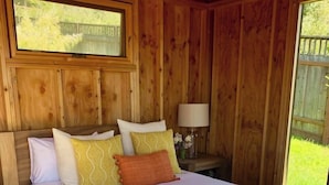 A wooden cabin bedroom with a bed, pillows, a nightstand, and a window. - Captain John's Hideaway (Stinson Beach)
